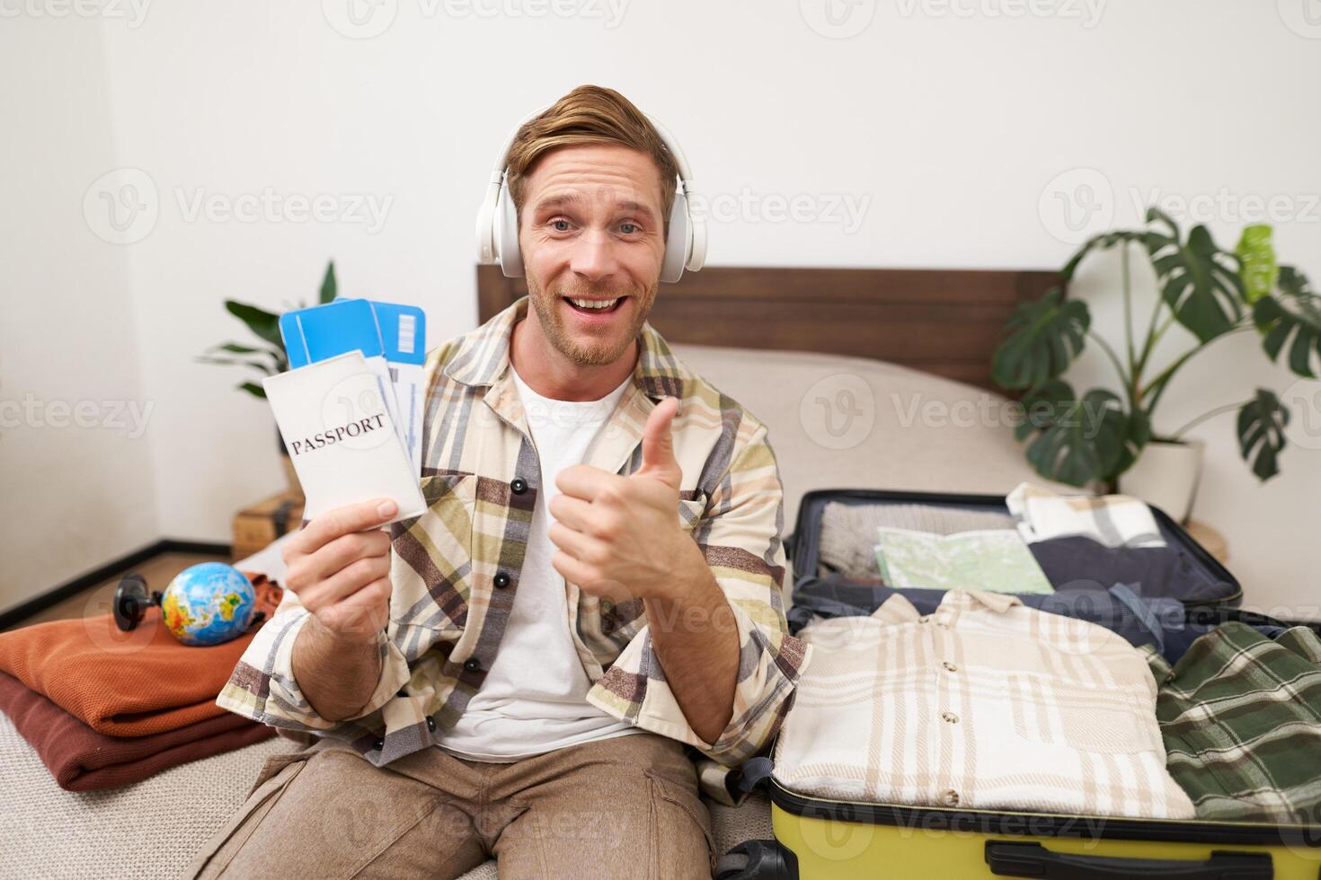 Portrait of smiling man, tourist shows two tickets and passport, goes on holiday, traveller makes thumb up in approval, like travel agency photo