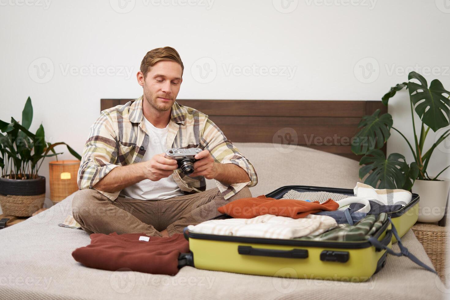 Portrait of happy, smiling young man sitting on bed, unpacking his suitcase after a holiday, tourist looking at his photo album on camera
