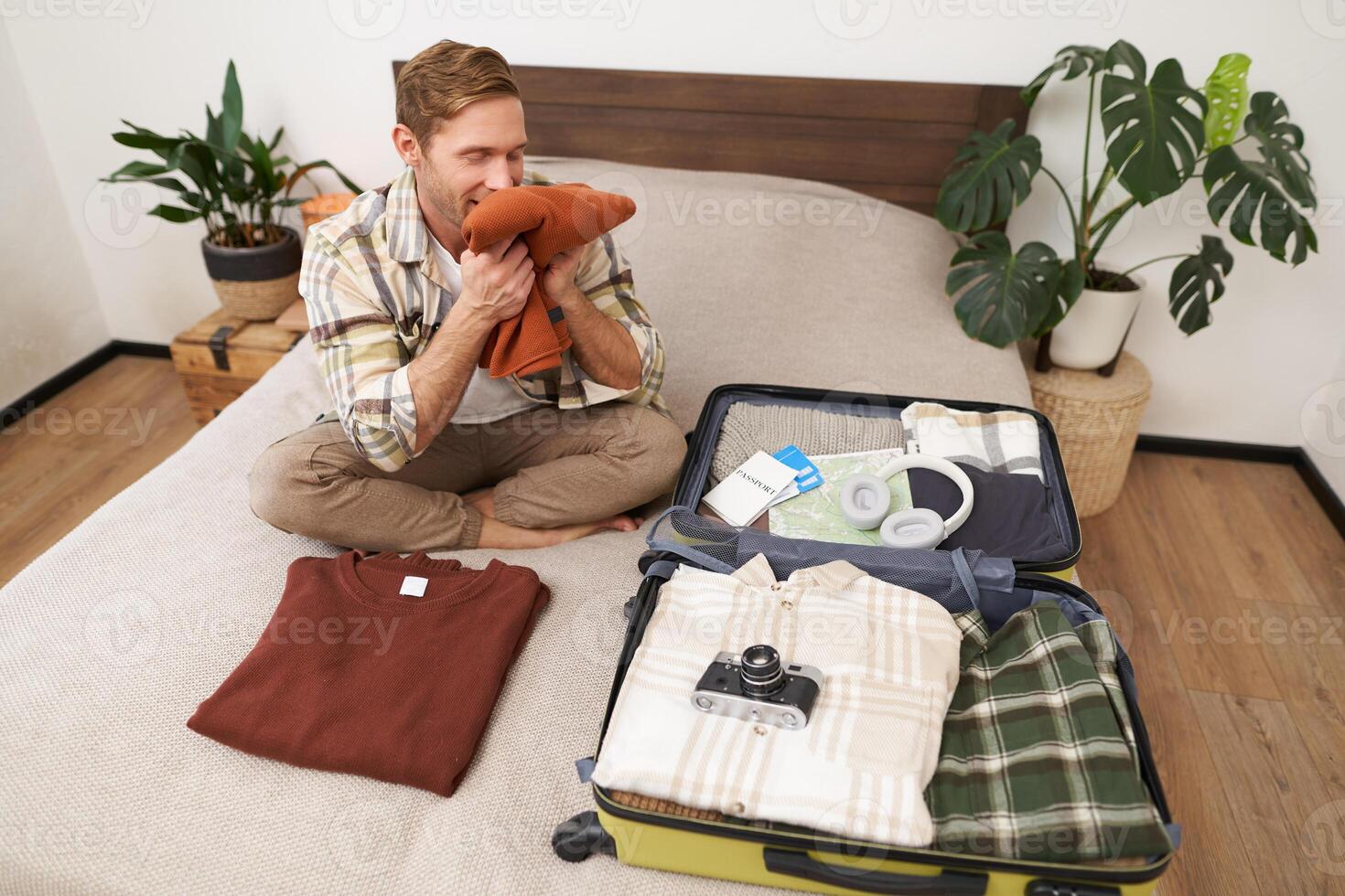 Portrait of happy young man, smells his clothes with pleased face, sits on bed with suitcase, goes on holiday, tries new detergent photo