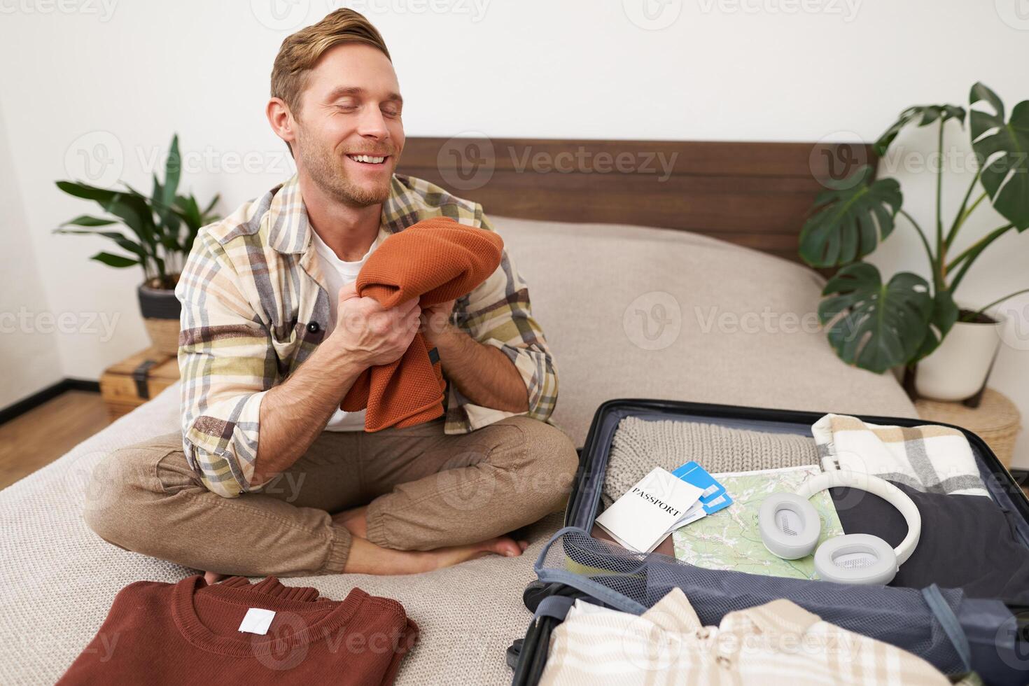 Portrait of smiling young man sits on bed with suitcase, packing for summer holiday, smelling clean and fresh clothes, goes on vacation photo