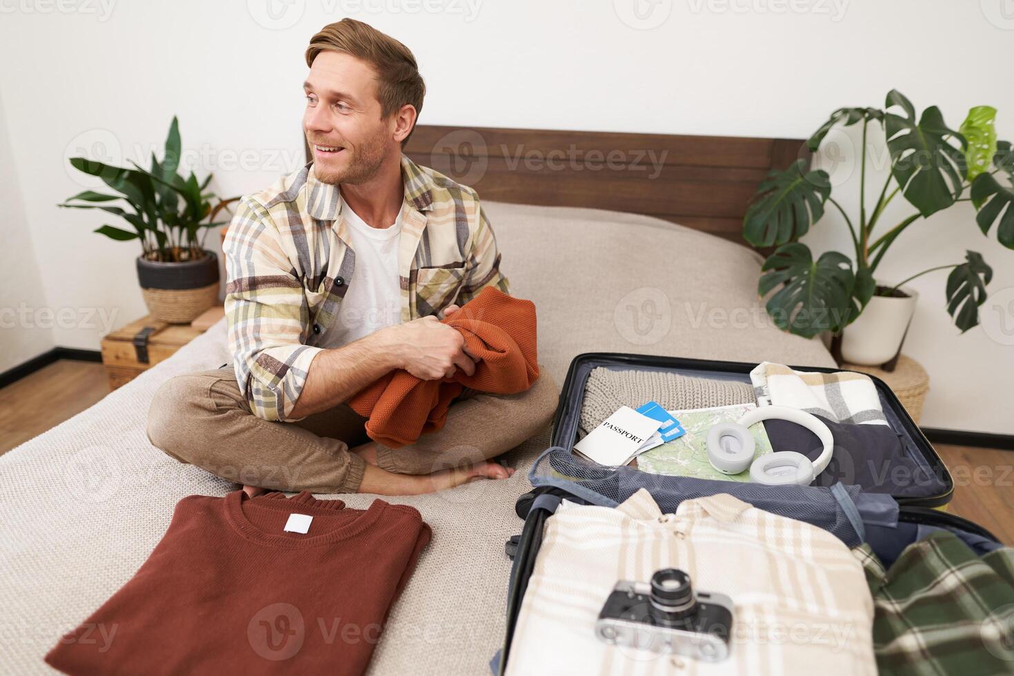 Image of young tourist, man unpacking his suitcase after summer vacation. Guy with luggage, sits on bed, packs clothes to go on holiday photo
