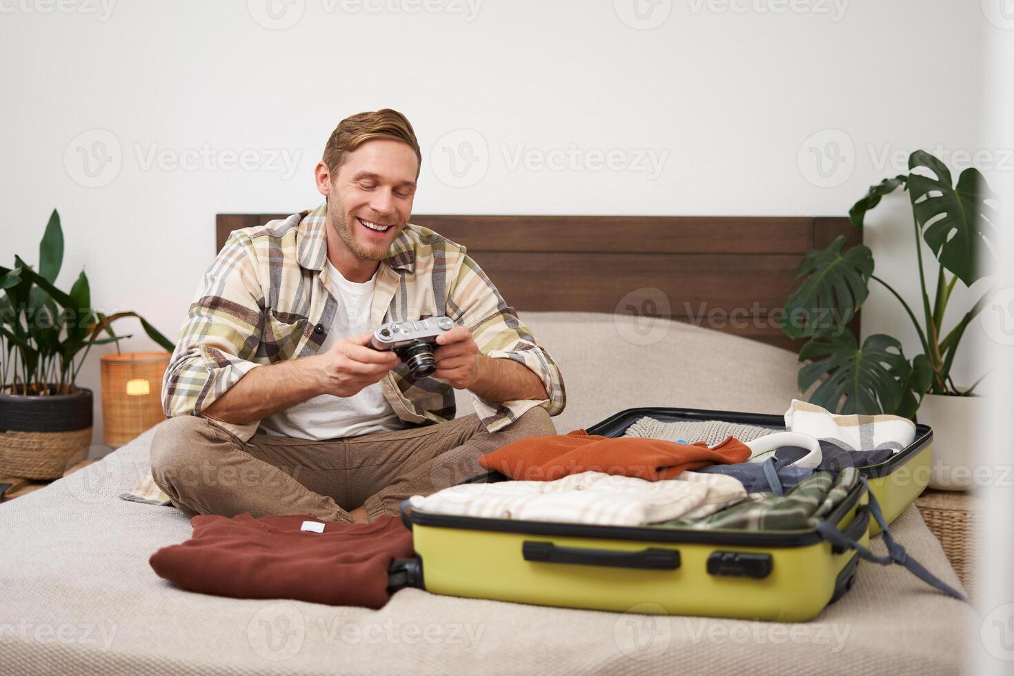 Portrait of happy, smiling young man sitting on bed, unpacking his suitcase after a holiday, tourist looking at his photo album on camera
