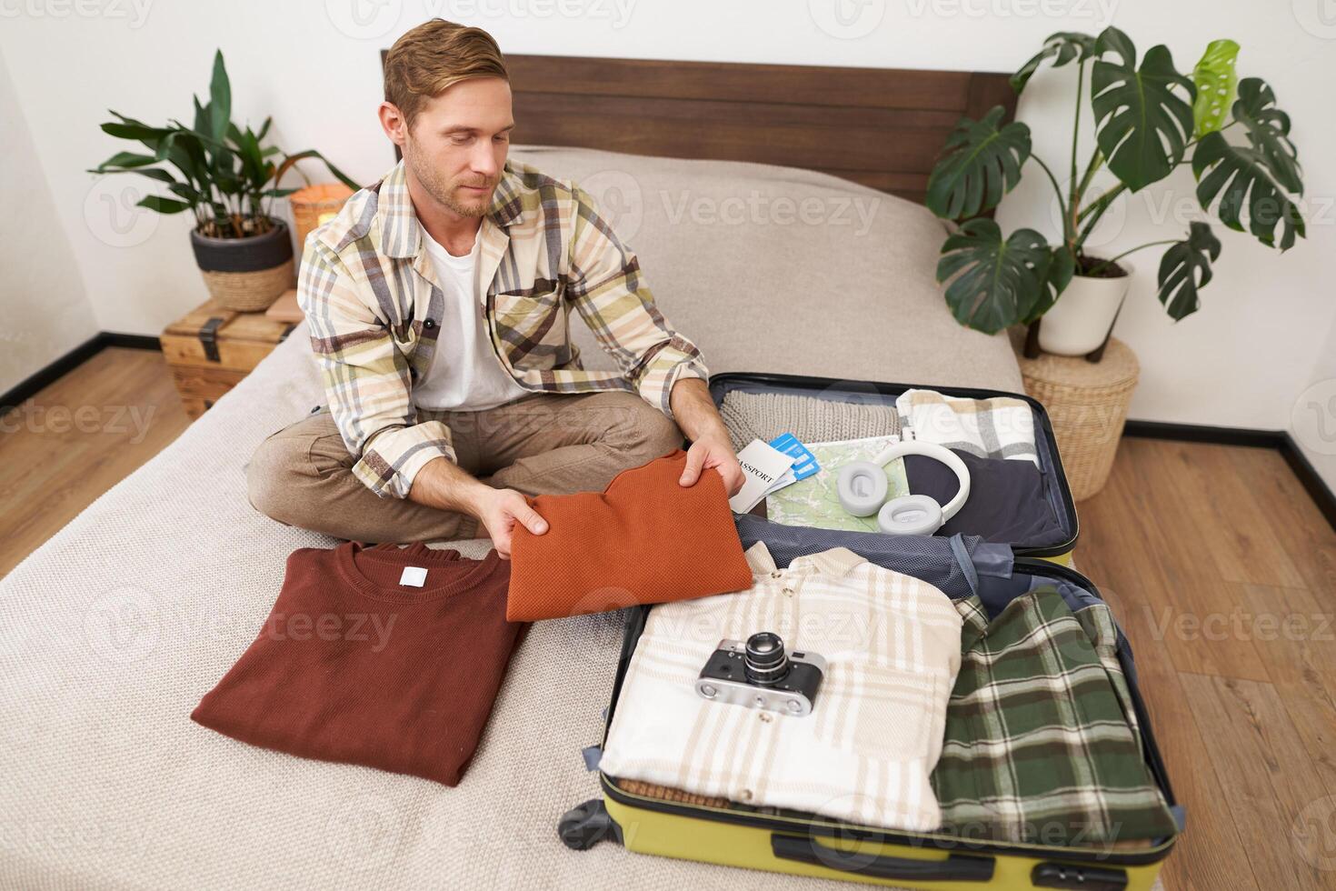 Image of young tourist, man unpacking his suitcase after summer vacation. Guy with luggage, sits on bed, packs clothes to go on holiday photo