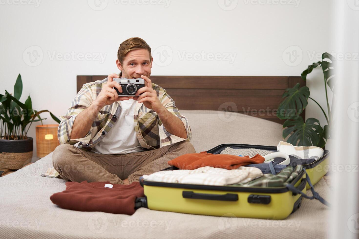Portrait of tourist, man sits with suitcase on bed, holds camera, packs suitcase with clothes, goes on holiday, prepares for summer vacation, tourist travels abroad photo