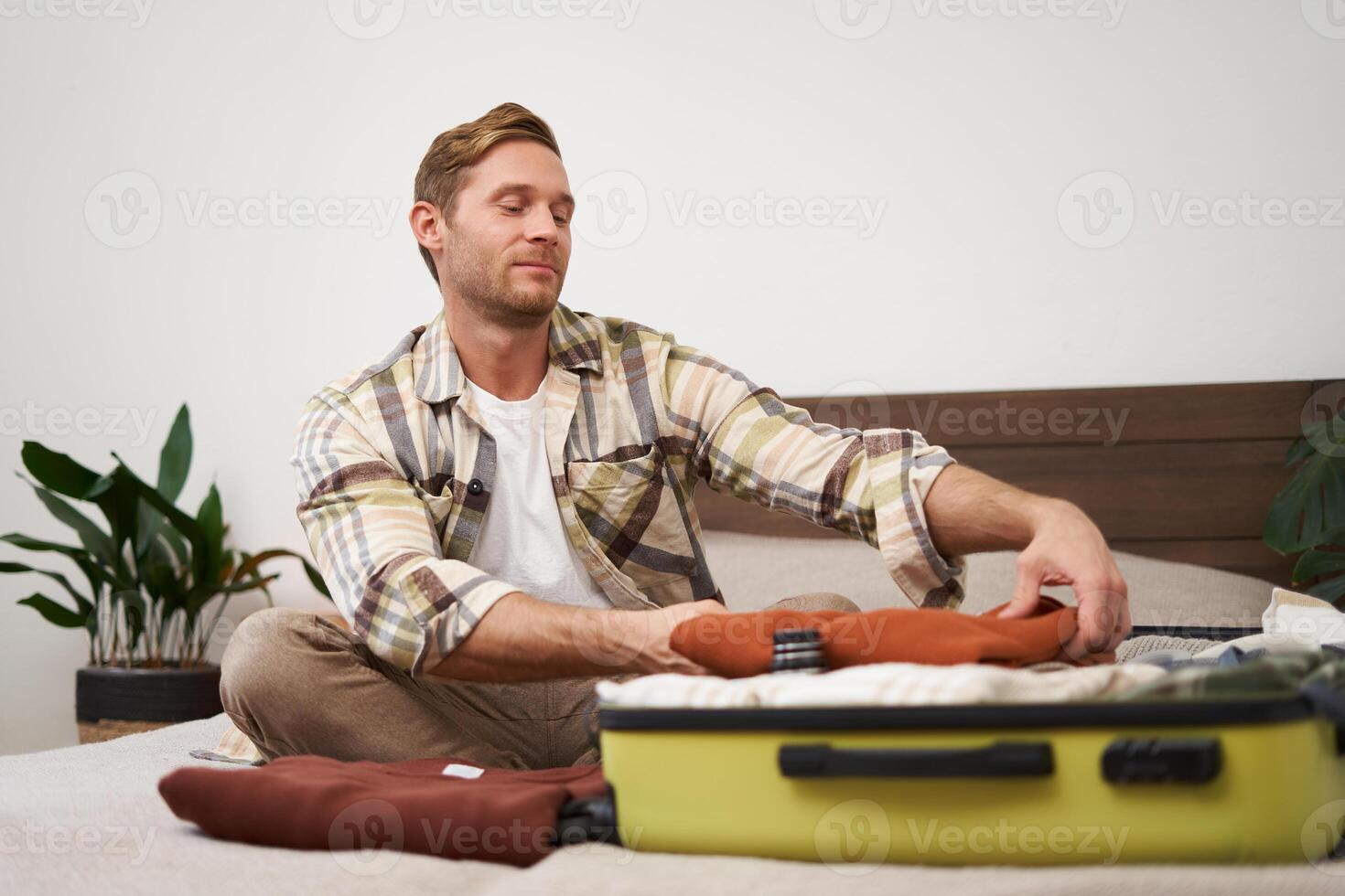 Portrait of handsome young man, tourist unpacking clothes in hotel room, staying in hostel on vacation, travelling, sitting with luggage on bed photo