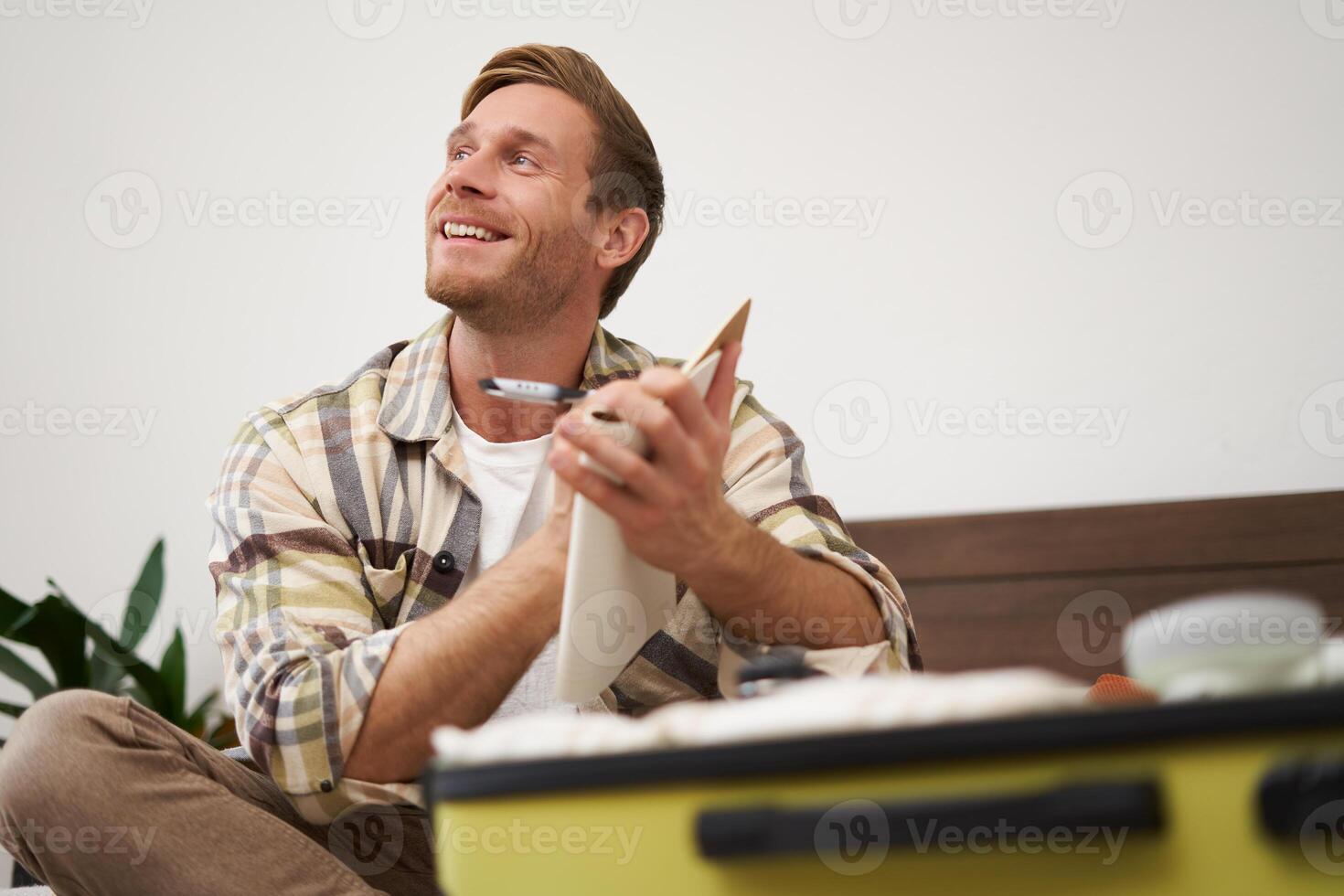 Close up portrait of smiling young man, writing down notes, holding pen and notebook, check-list of items in suitcase, going on holiday photo