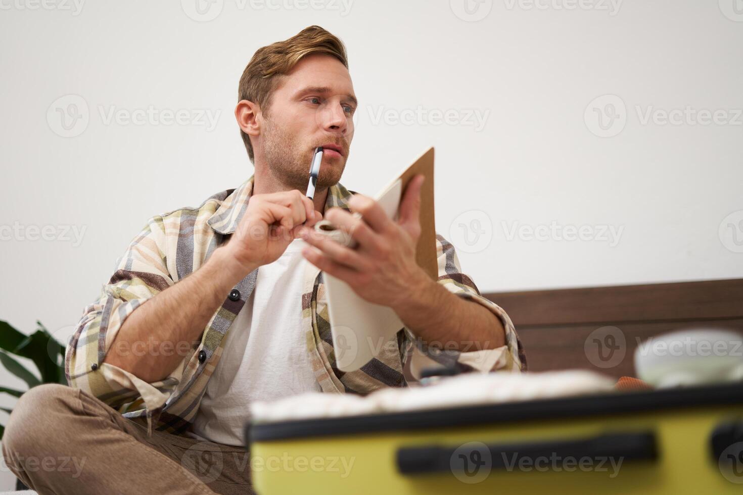 Portrait of young man, tourist looking at his notes, check-list of items to bring on vacation, packing clothes in suitcase, going on holiday photo