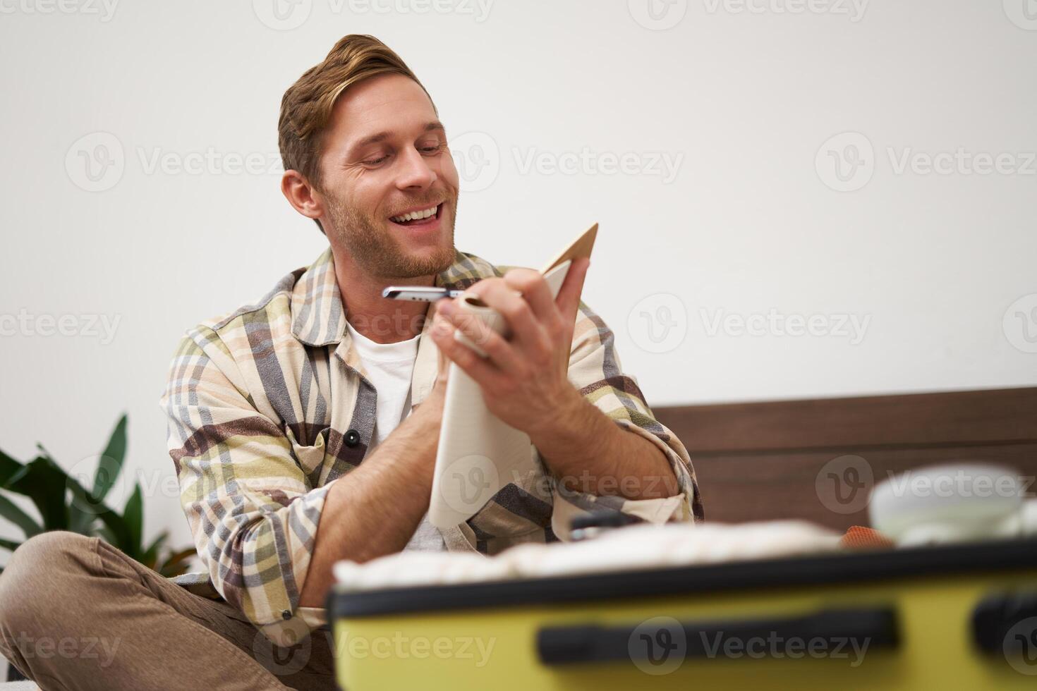 Portrait of young man, tourist looking at his notes, check-list of items to bring on vacation, packing clothes in suitcase, going on holiday photo