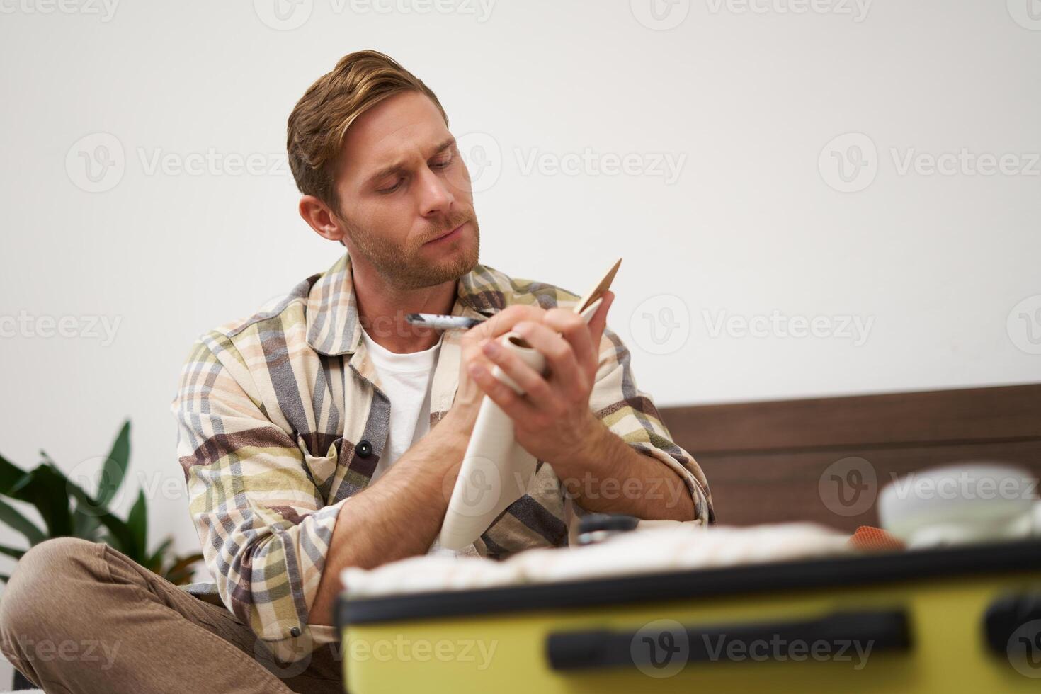 Portrait of tourist packing suitcase, going through check-list, writing down items to take with him in luggage, making notes in notebook, sitting on bed photo