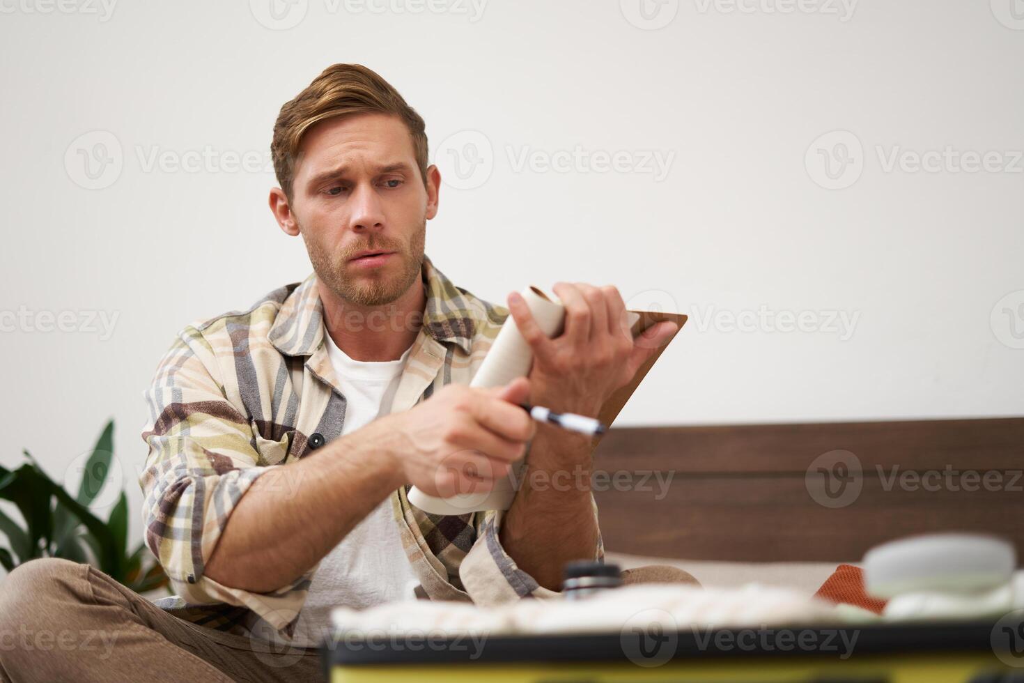 Portrait of young man looking serious at his suitcase with clothes, going through the check-list, holding notebook with notes, choosing what to bring on holiday. Tourism concept photo