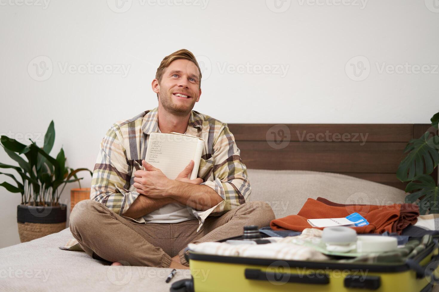 Portrait of thoughtful young man, tourist planning his vacation, sitting with open suitcase on bed, making notes, writing to do check list in notebook photo