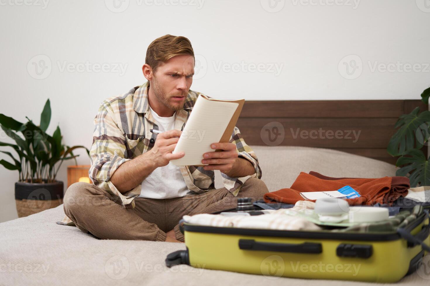 Portrait of confused man, looks serious at his notes, reading something and frowning from disapproval, packing or unpacking suitcase in hotel room, sitting on bed photo