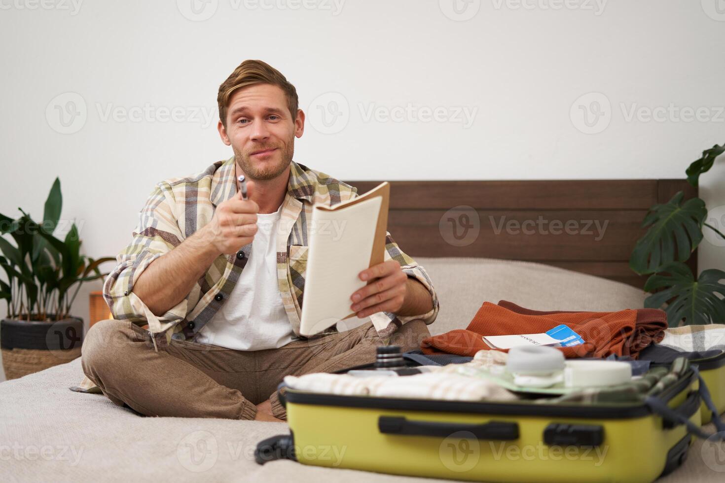 Portrait of man with suitcase, sitting and packing things for holiday, checking list of items for vacation, writing down, making notes in notebook photo