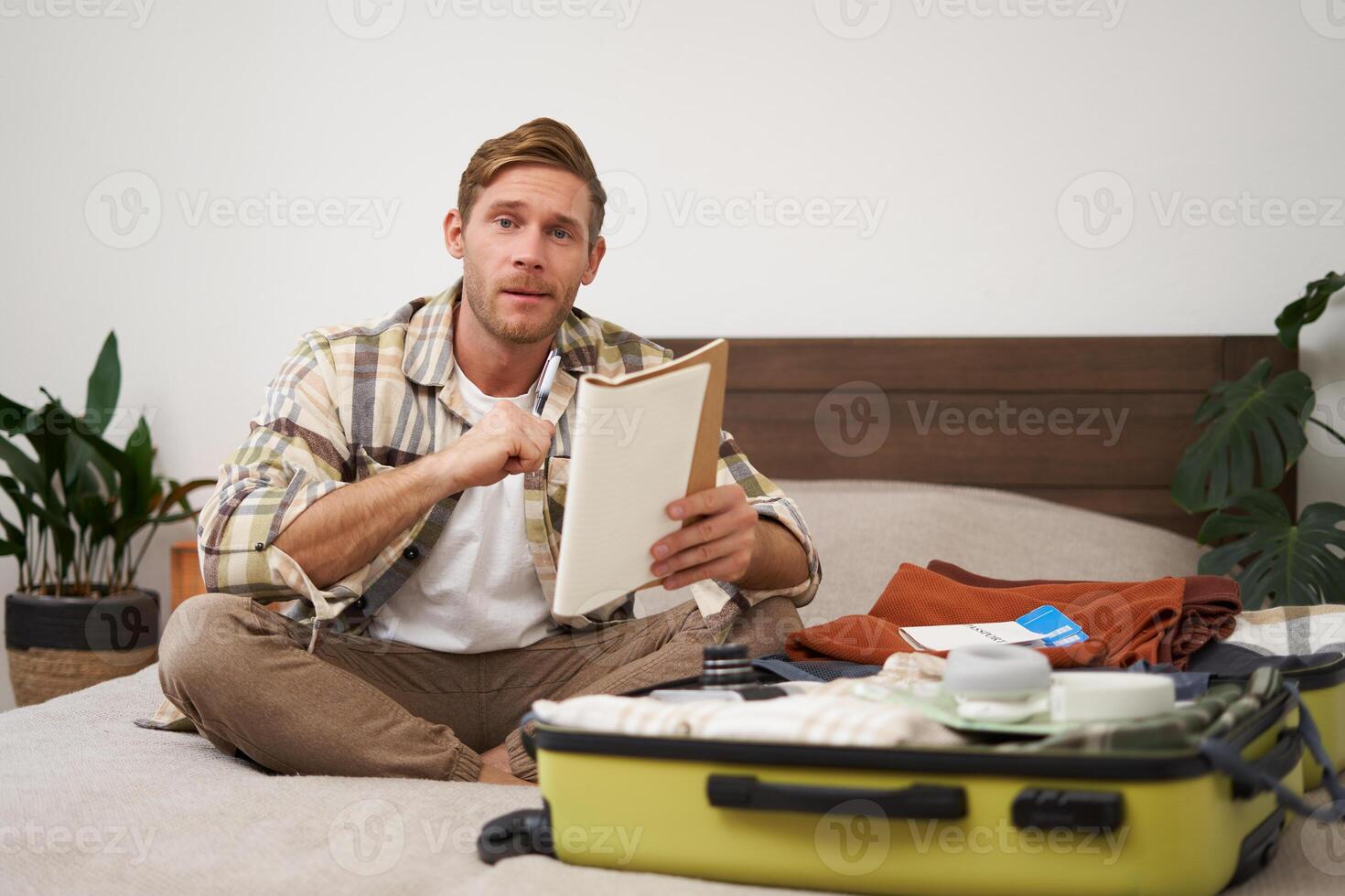 Portrait of young man, looking happy, going on holiday trip, travelling, holding notebooks with list of items, packing suitcase while sitting on bed photo
