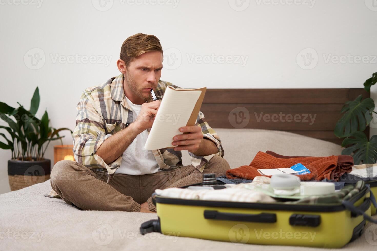 Portrait of angry man, packing suitcase, looking frustrated at notebook with check list of items for holiday, preparing to go on vacation photo
