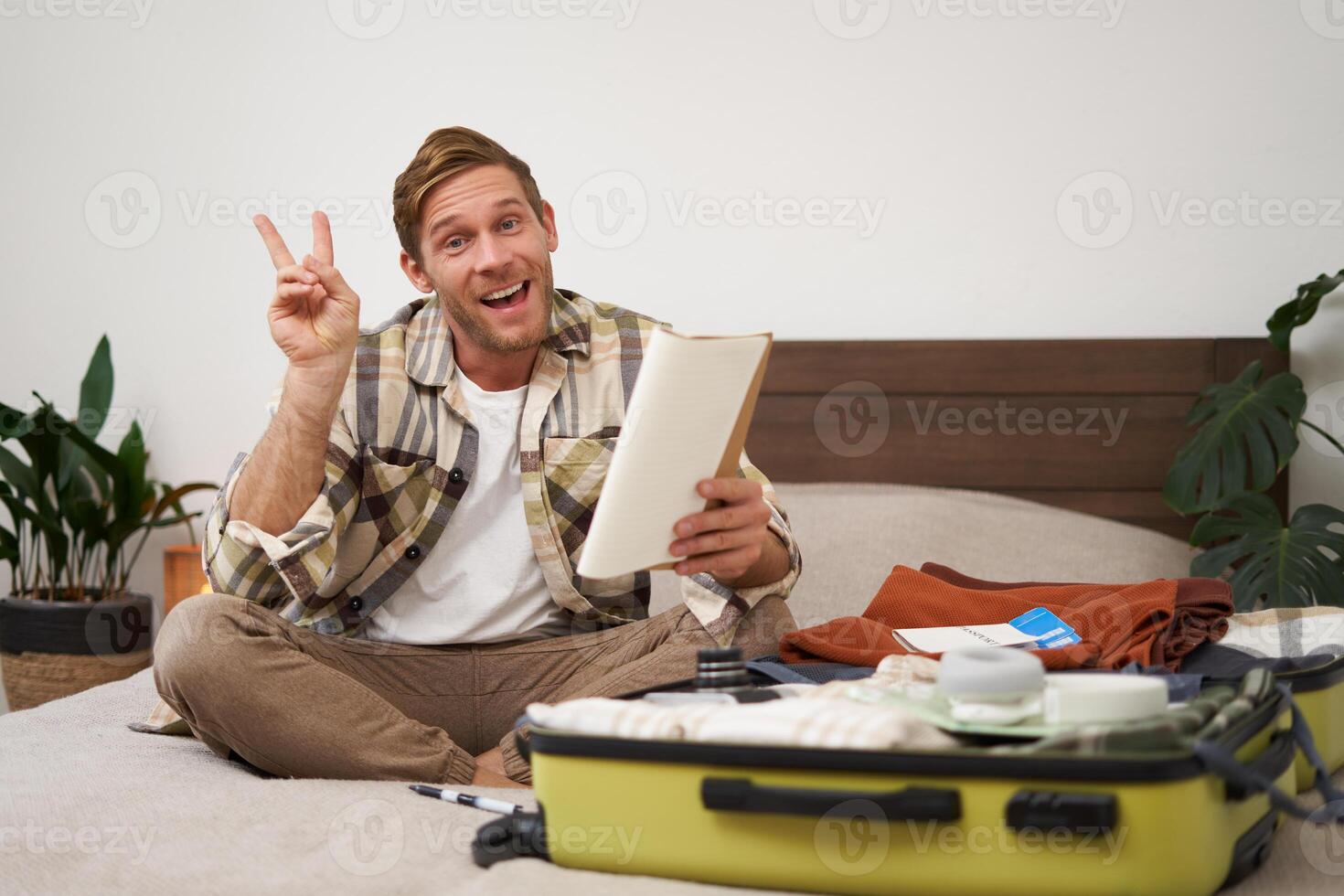 Portrait of young man, looking happy, going on holiday trip, travelling, holding notebooks with list of items, packing suitcase while sitting on bed photo