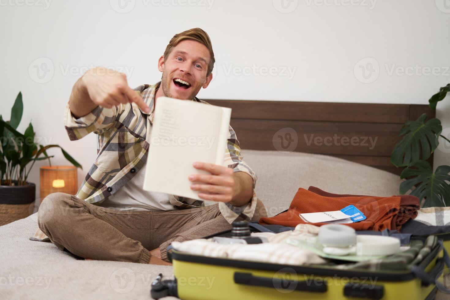 Portrait of young man pointing at his list of items, packing clothes on vacation, showing notebook, sitting near opened suitcase photo