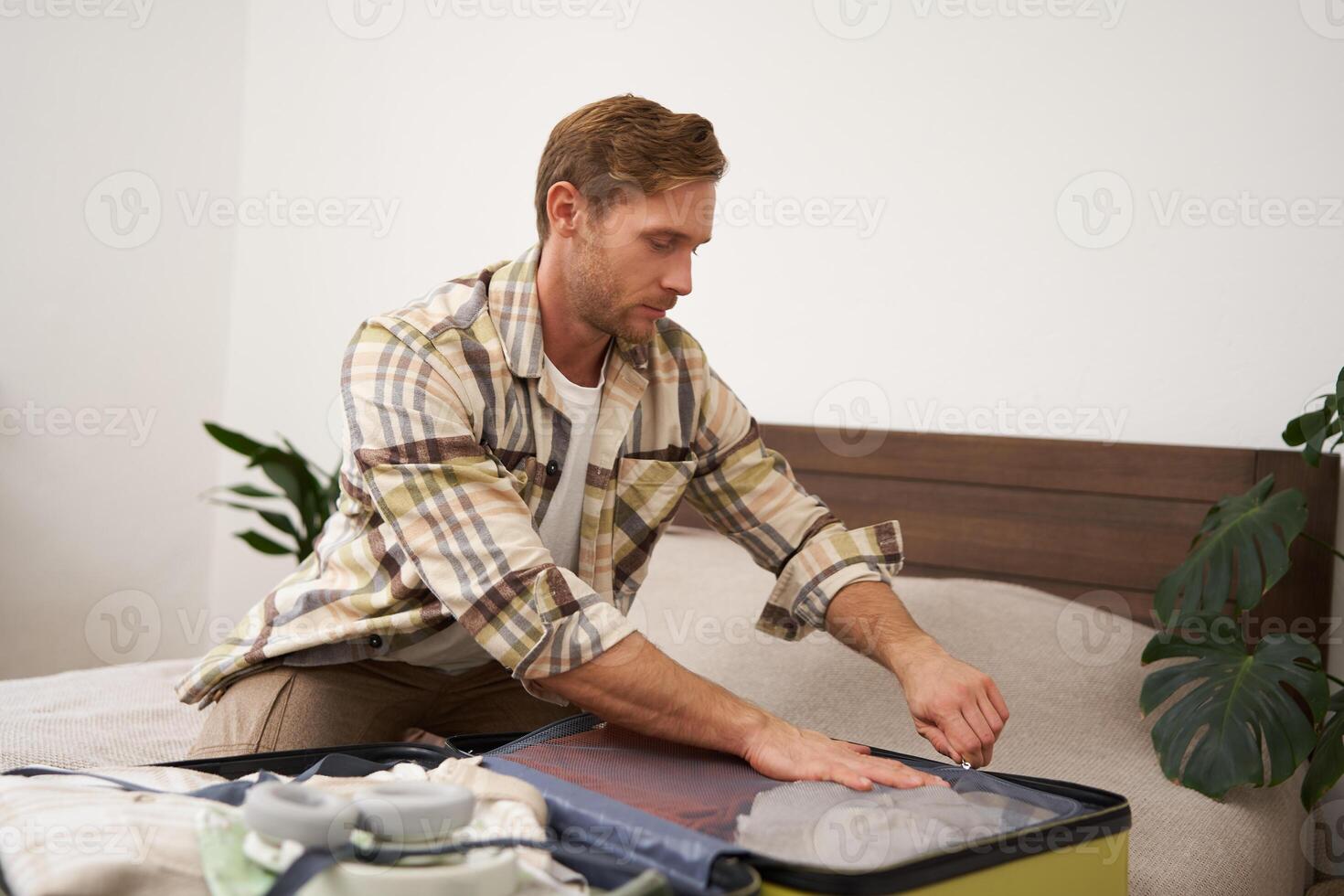 Portrait of young man, tourist going travelling abroad, putting clothes in suitcase, closing his luggage, sitting on bed, getting ready for holiday photo