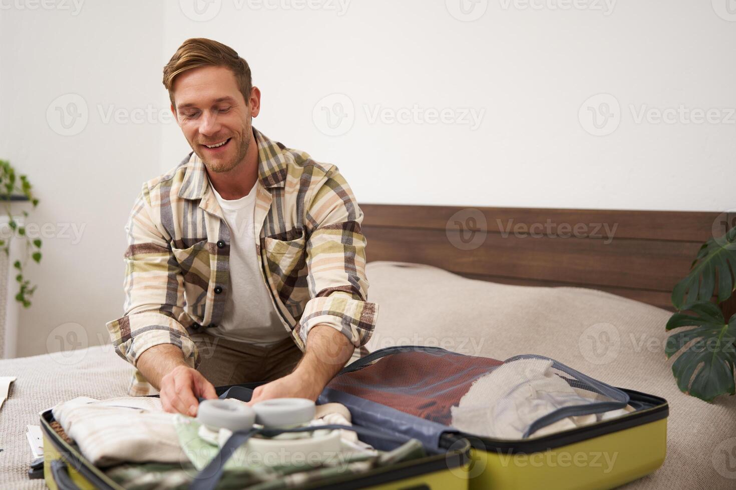 Portrait of young man, tourist going travelling abroad, putting clothes in suitcase, closing his luggage, sitting on bed, getting ready for holiday photo