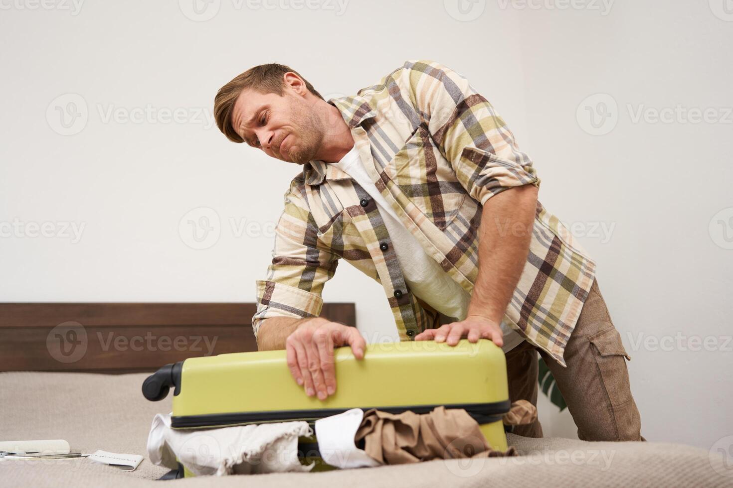 Side view of young man pushing on suitcase, trying to close it, has lots of clothes sticking out of luggage, going on long holiday, packing for vacation photo