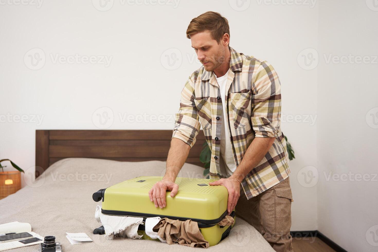 Image of young man, tourist struggling to close his suitcase full of clothes, pushing on bag, trying to press items inside luggage to shut the zipper, packing for travelling abroad, going on holiday photo