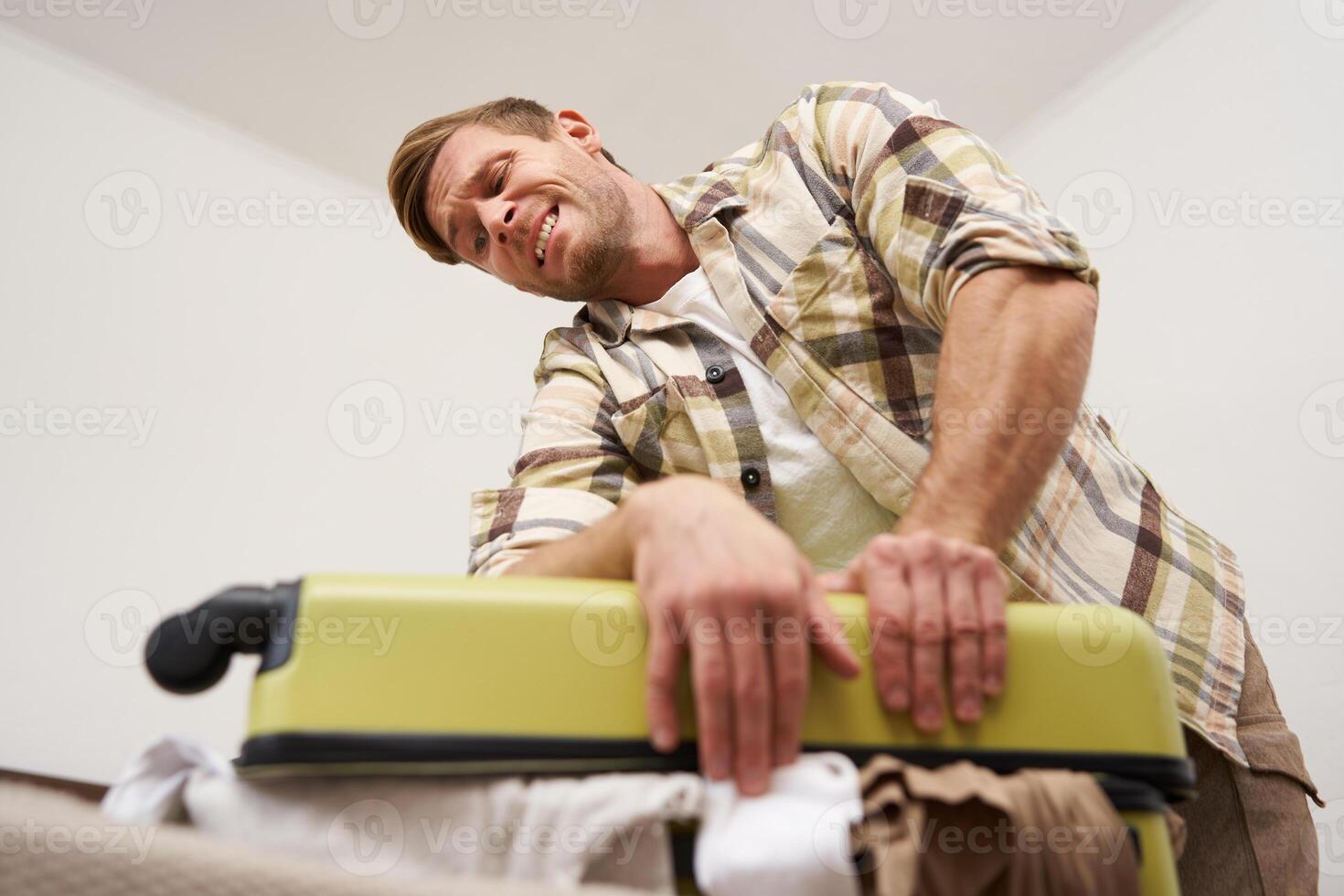 Side view of young man pushing on suitcase, trying to close it, has lots of clothes sticking out of luggage, going on long holiday, packing for vacation photo