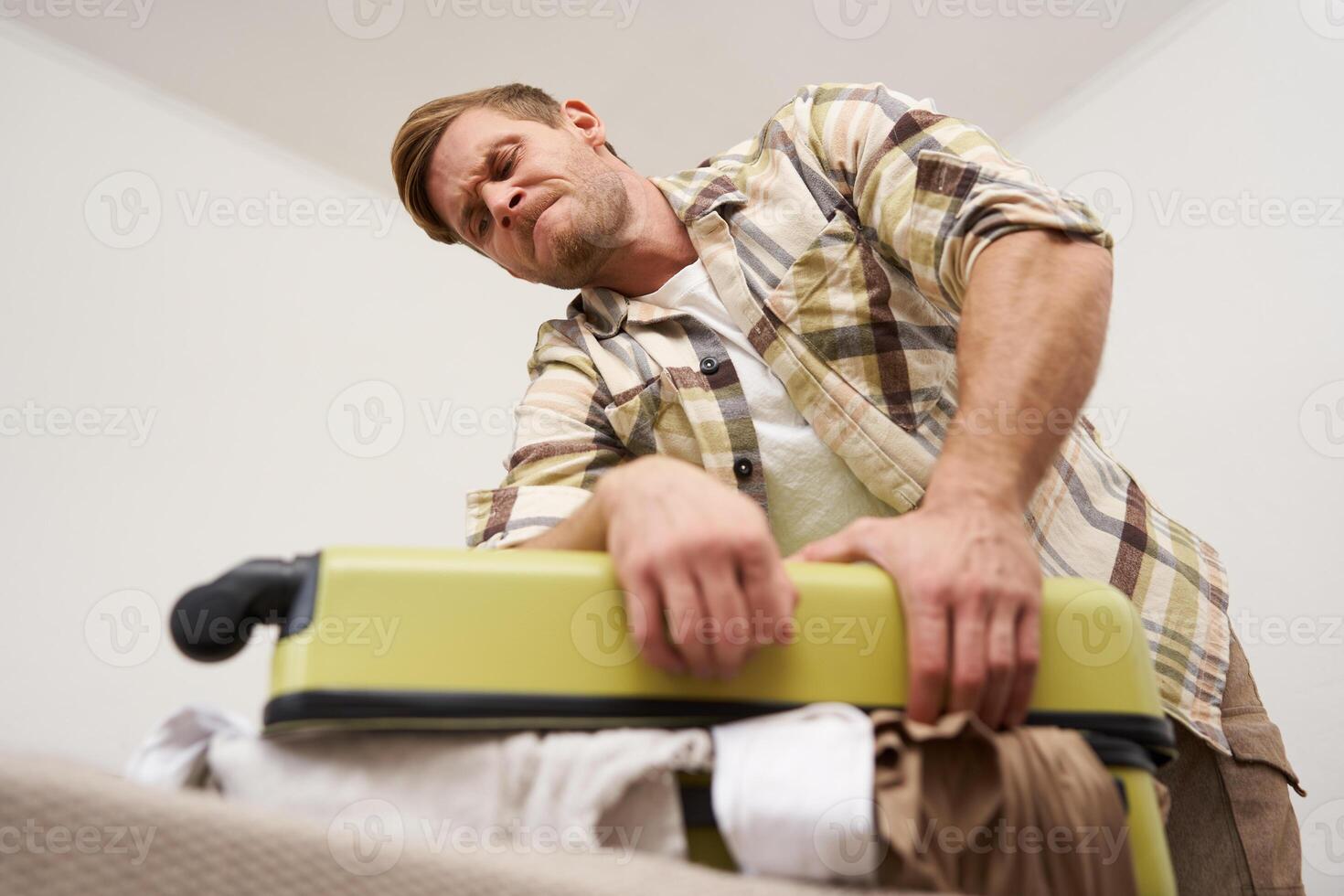 Image of young man, tourist struggling to close his suitcase full of clothes, pushing on bag, trying to press items inside luggage to shut the zipper, packing for travelling abroad, going on holiday photo