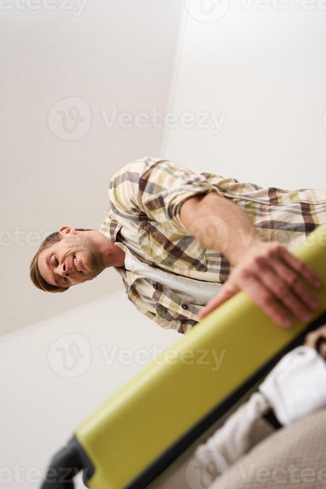Vertical shot of man trying to press suitcase and close zipper, packing clothes for vacation. Tourist going on holiday, preparing for travelling abroad photo
