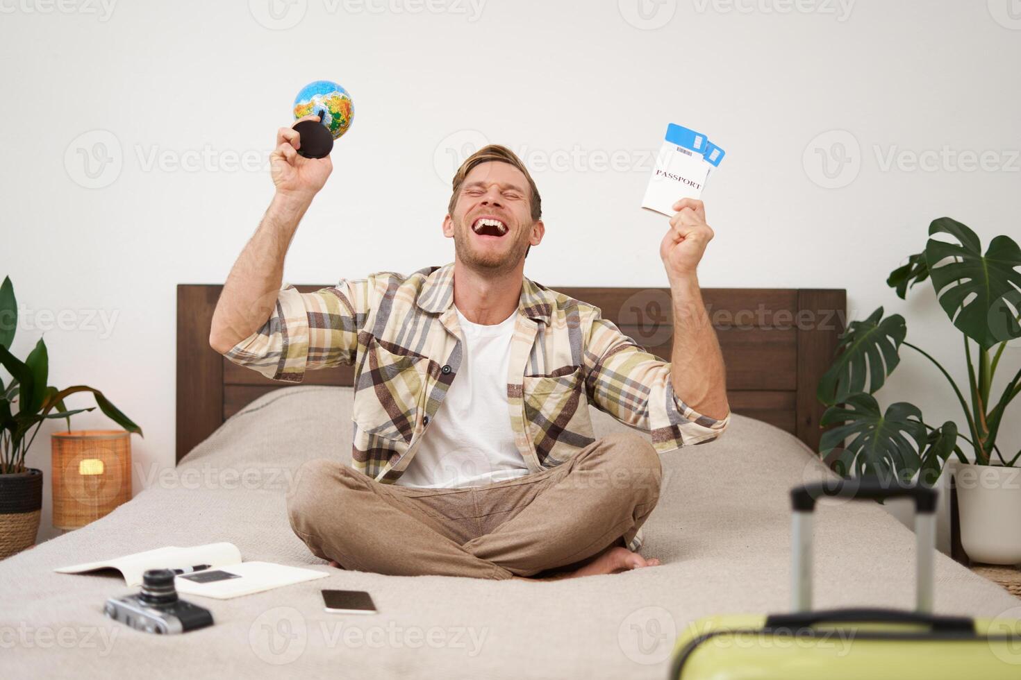 Portrait of happy guy, tourist goes on vacation abroad, holds globe and plane ticket, excited about his holiday, sits on bed with smiling, enthusiastic face expression photo