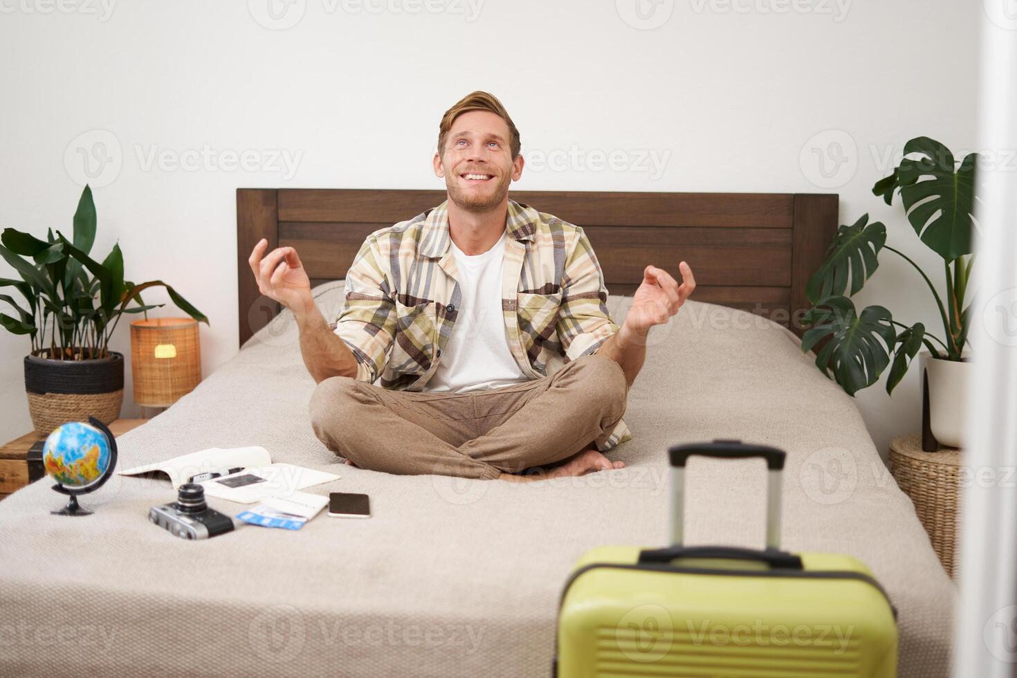Close up portrait of meditating tourist, man sits with suitcase in bedroom, practice yoga, relaxing, finding nirvana photo