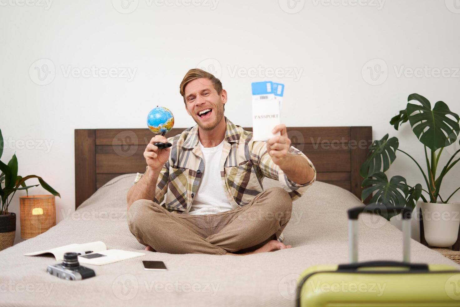 Portrait of happy guy, tourist goes on vacation abroad, holds globe and plane ticket, excited about his holiday, sits on bed with smiling, enthusiastic face expression photo