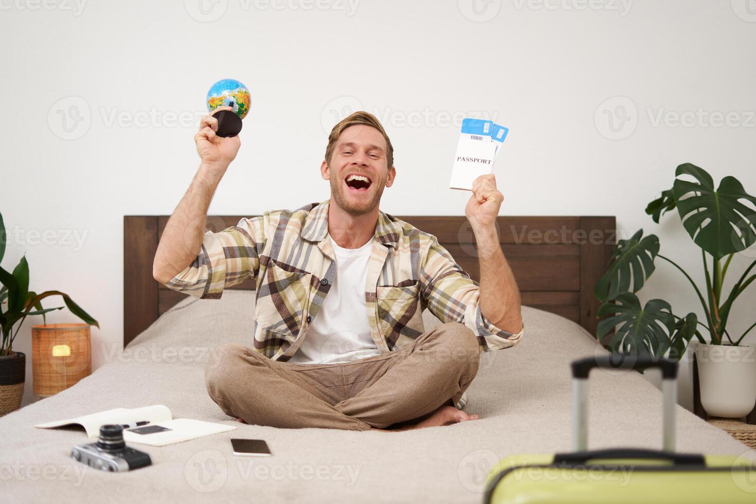 Portrait of cheerful, happy tourist, man sitting with plane tickets and a globe, packed suitcase for travelling around the world, going on vacation, excited about his holiday photo