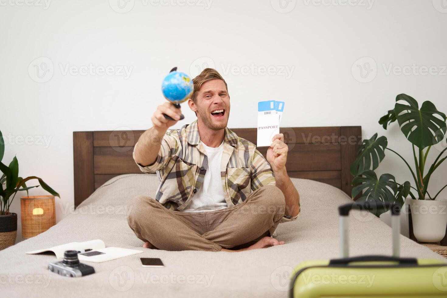Excited, handsome young man sits on bed, shows globe and tickets, has suitcase, camera packed for holiday, goes on vacation, happy to go on holiday photo