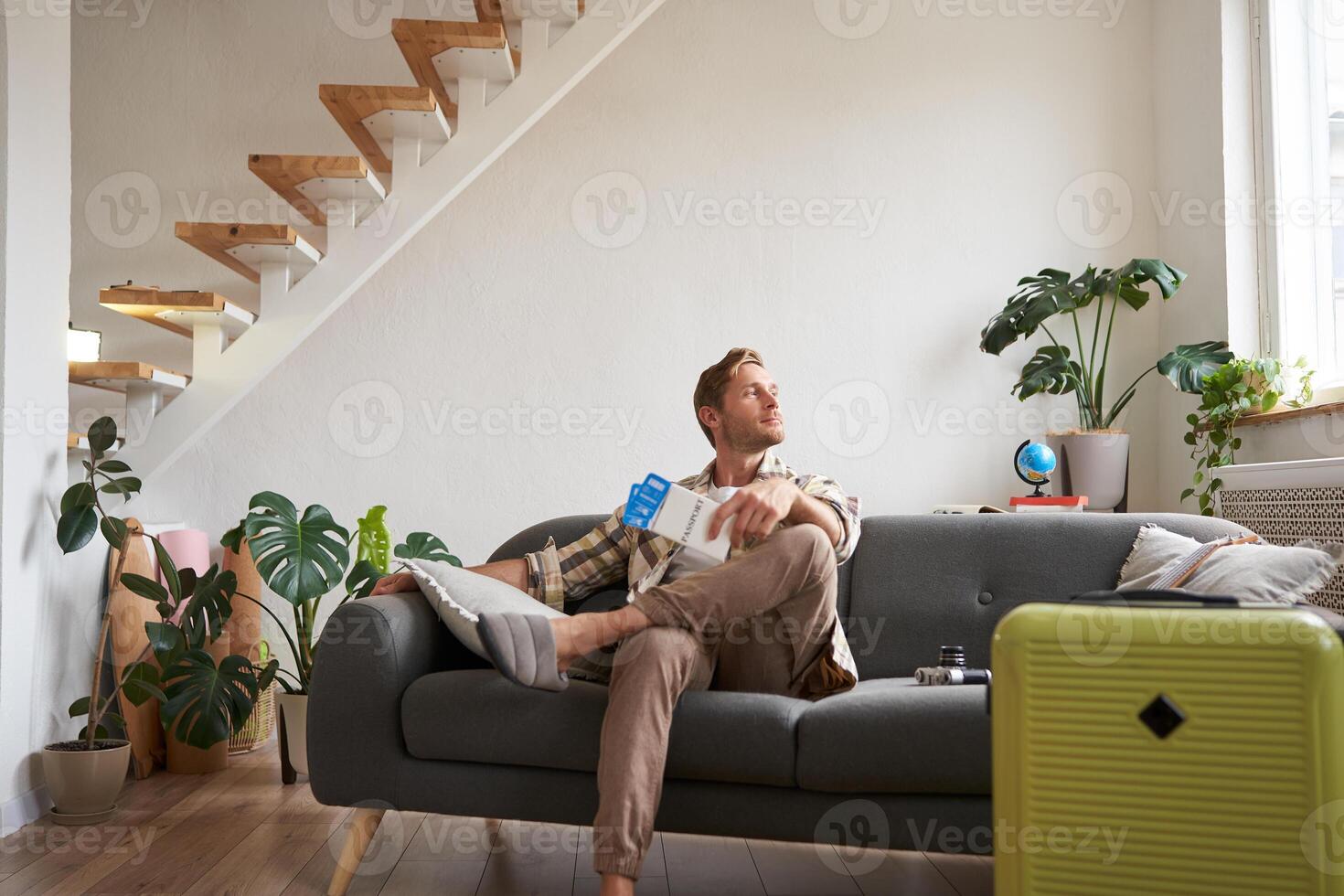 Portrait of handsome young man, sits with suitcase in living room, holds two plane tickets, going on vacation, waiting for taxi, looking outside window photo