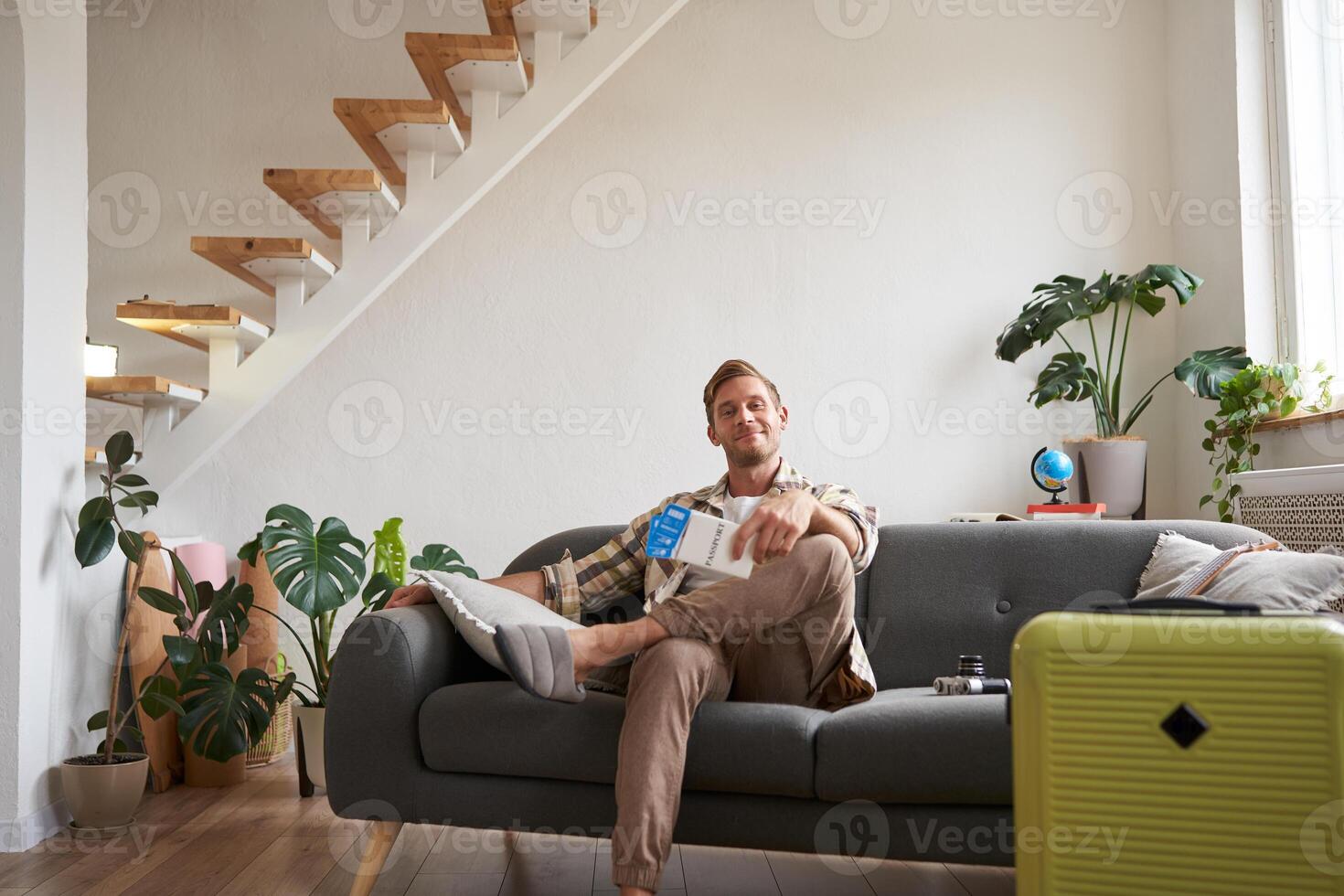 Portrait of handsome young man, bought tickets for plane, going on holiday, sitting on sofa with packed suitcase, waiting for taxi to airport photo