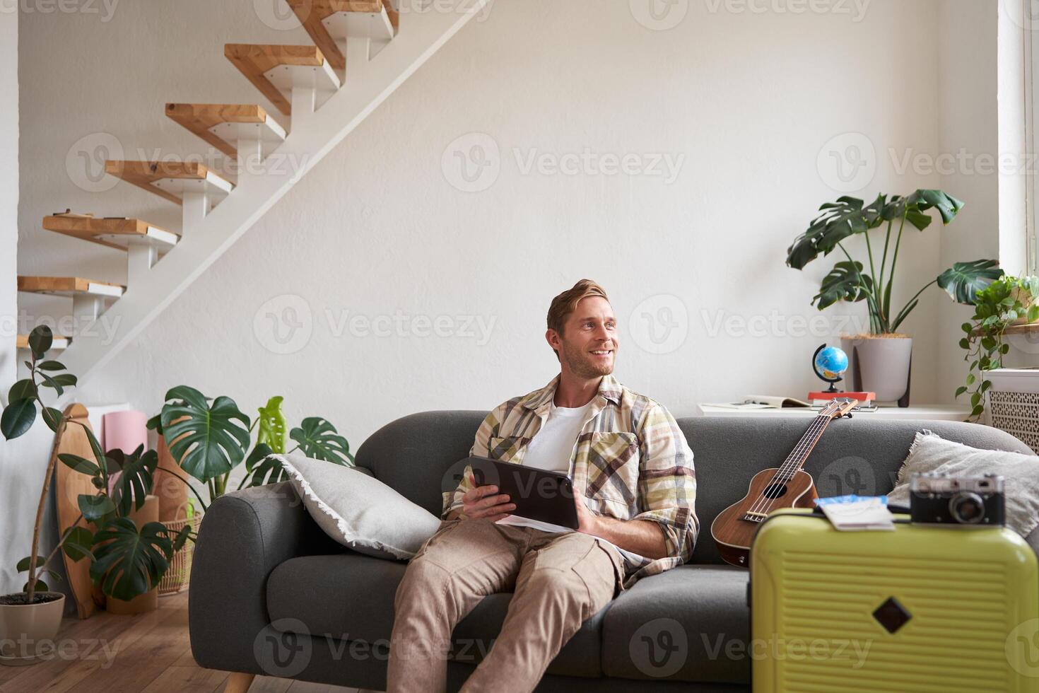 Image of young man, tourist goes on holiday, packed suitcase, sits with digital tablet on sofa, reads, looks at his map, books hotel for business trip photo