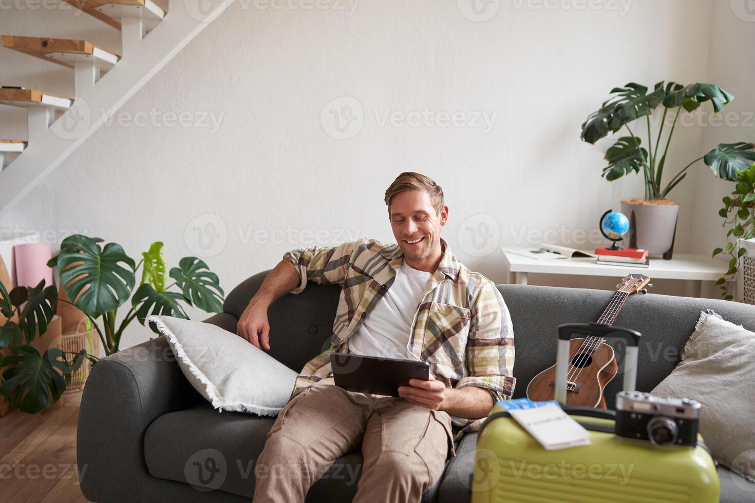 Image of young man with tablet, sitting in living room with suitcase, reading, studying the route, preparing for a vacation photo