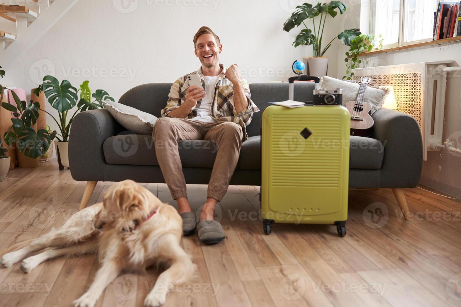 Image of excited man, tourist with a dog, sits in living room with packed suitcase and travel camera, guy saying yes and celebrating, pleased with prices online, booking pet-friendly hotel photo