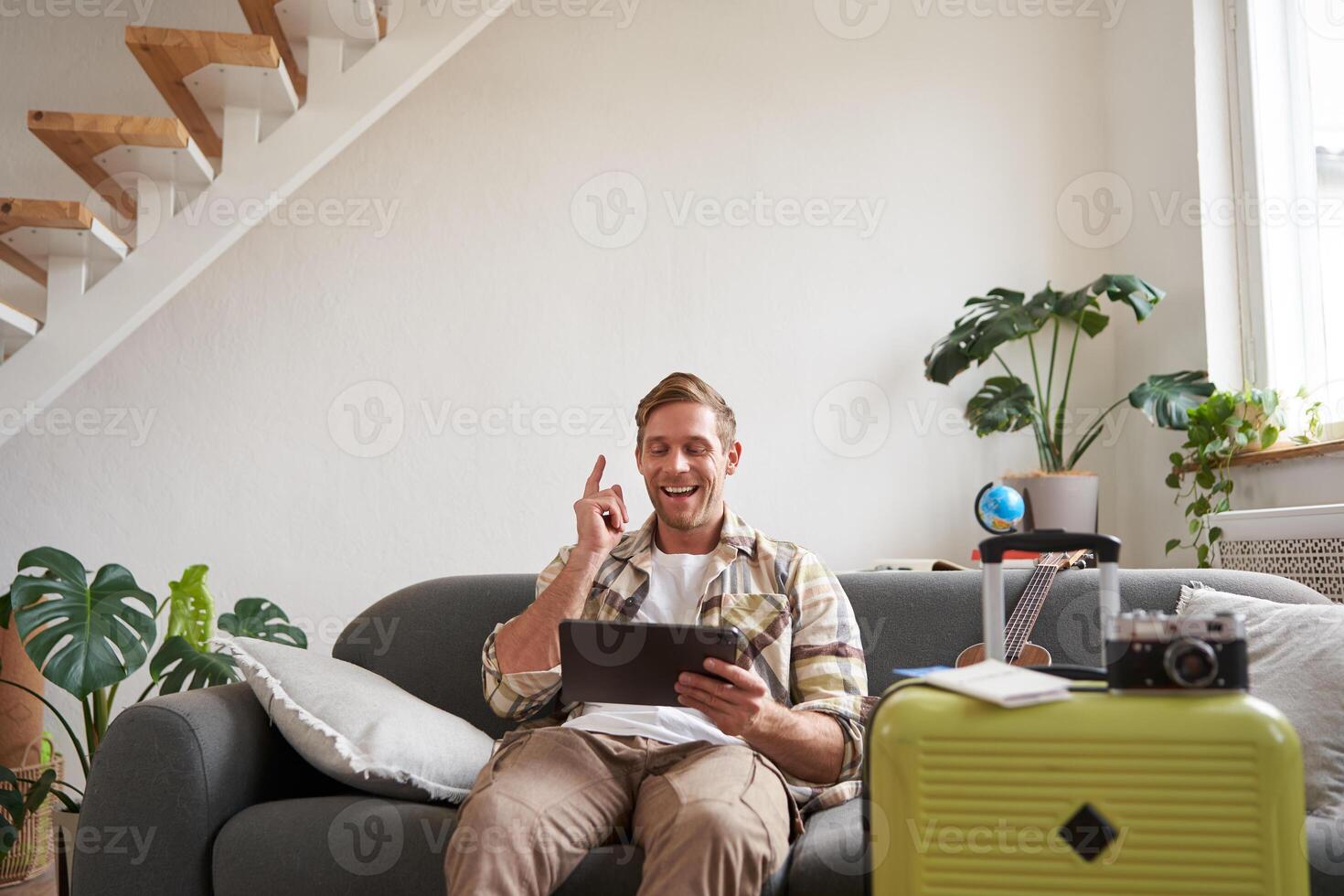 Image of young man, tourist goes on holiday, packed suitcase, sits with digital tablet on sofa, reads, looks at his map, books hotel for business trip photo