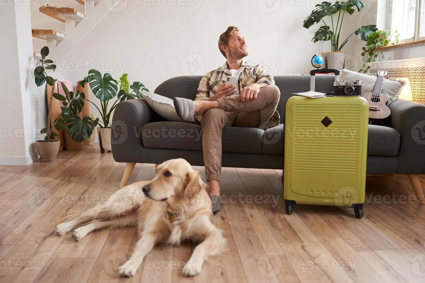 Portrait of happy, handsome young man with suitcase, sitting with his dog in living room, prepared to go on vacation, going on holiday photo