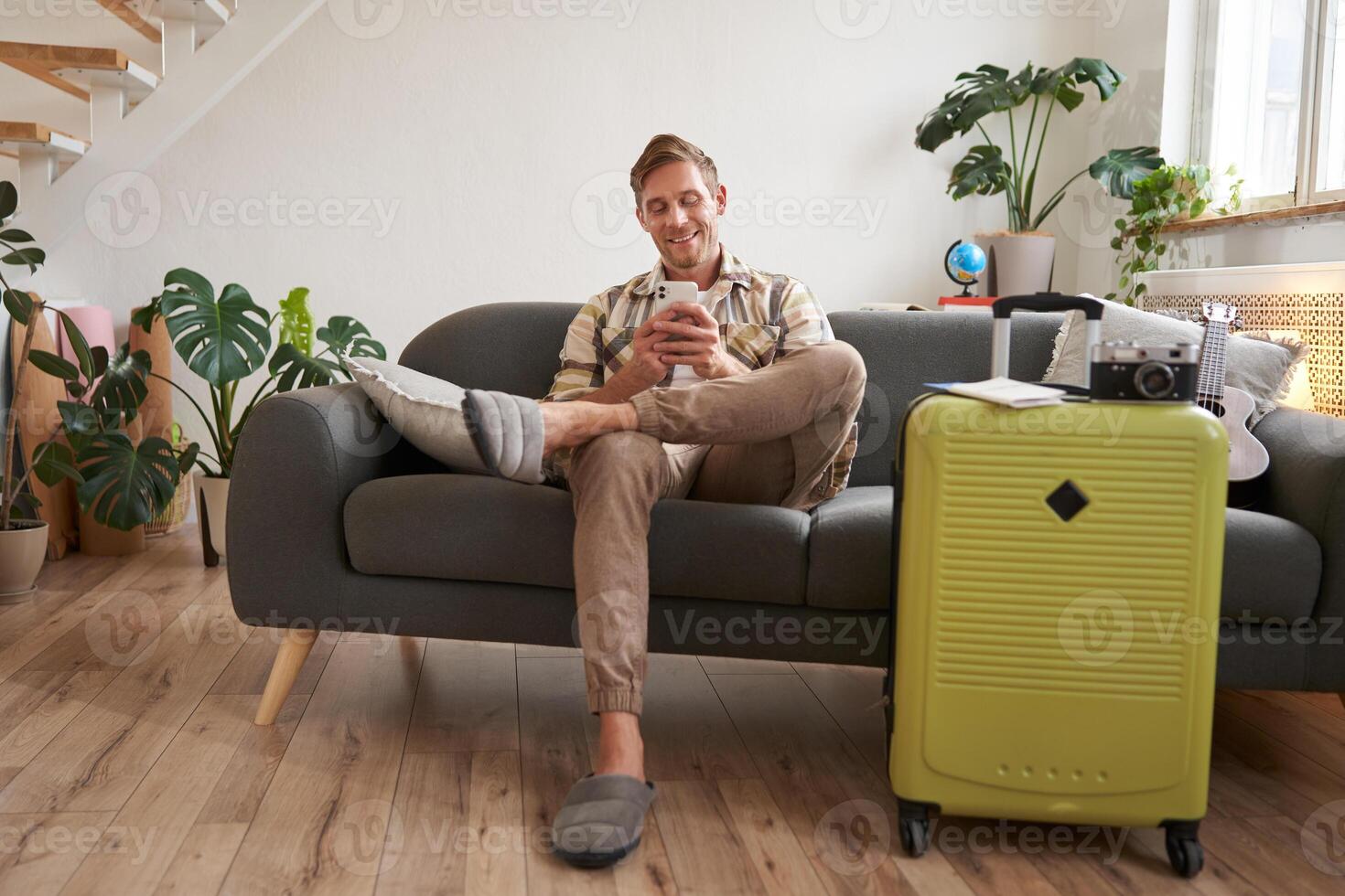 Handsome young man sits on sofa with suitcase, holds smartphone, books hostel or holiday vacation, chats on mobile phone photo