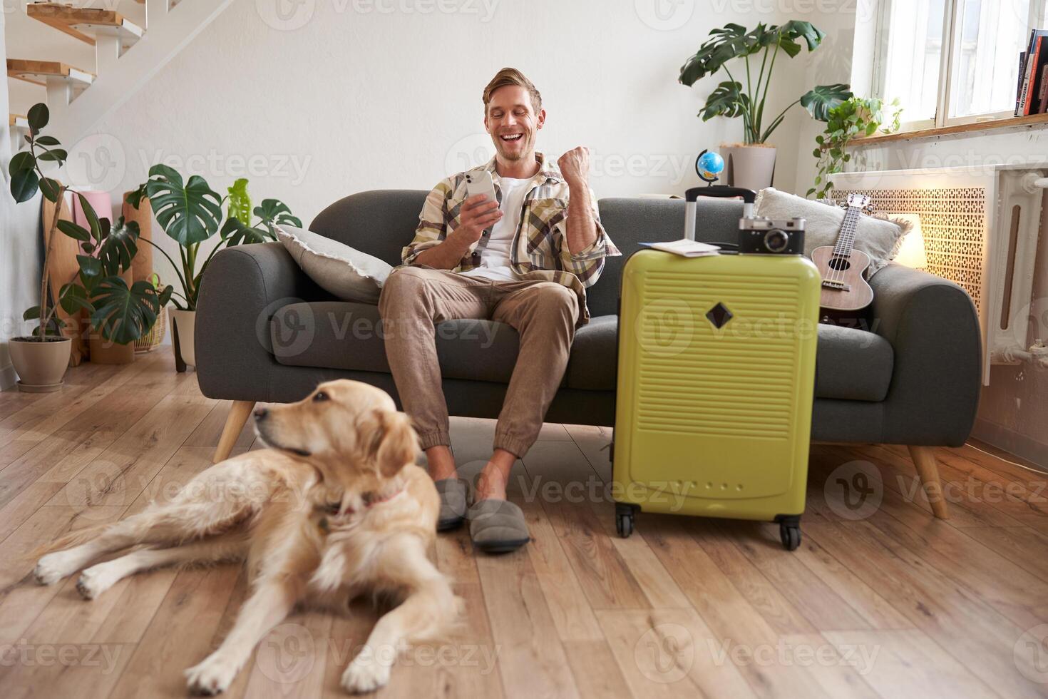 Happy man with his dog, sits in living room with suitcase, looks at smartphone app with excited face, pleased with booked trip photo