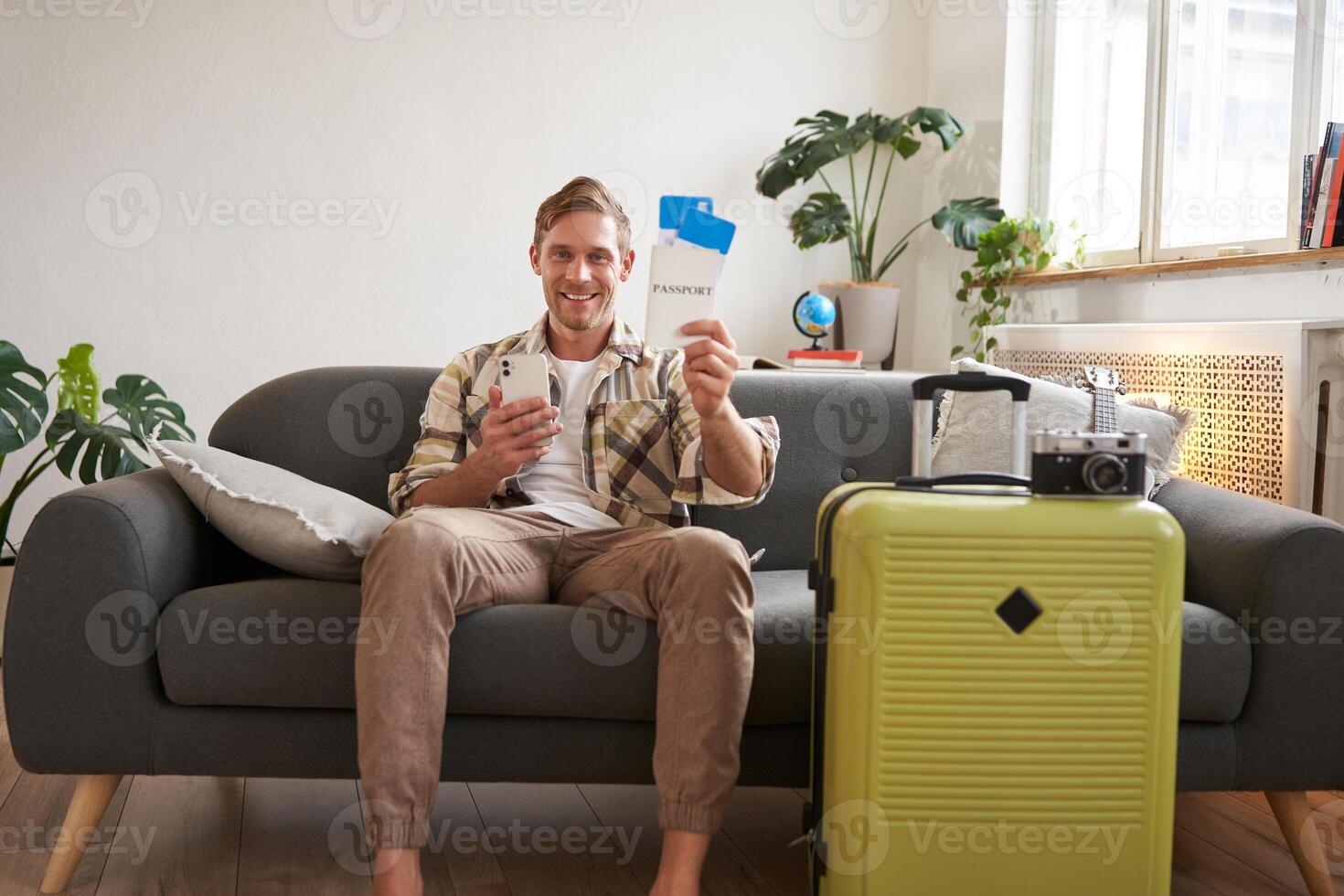 Image of handsome male model, tourist with suitcase, shows his two tickets on plane, uses mobile phone app, sits on sofa photo