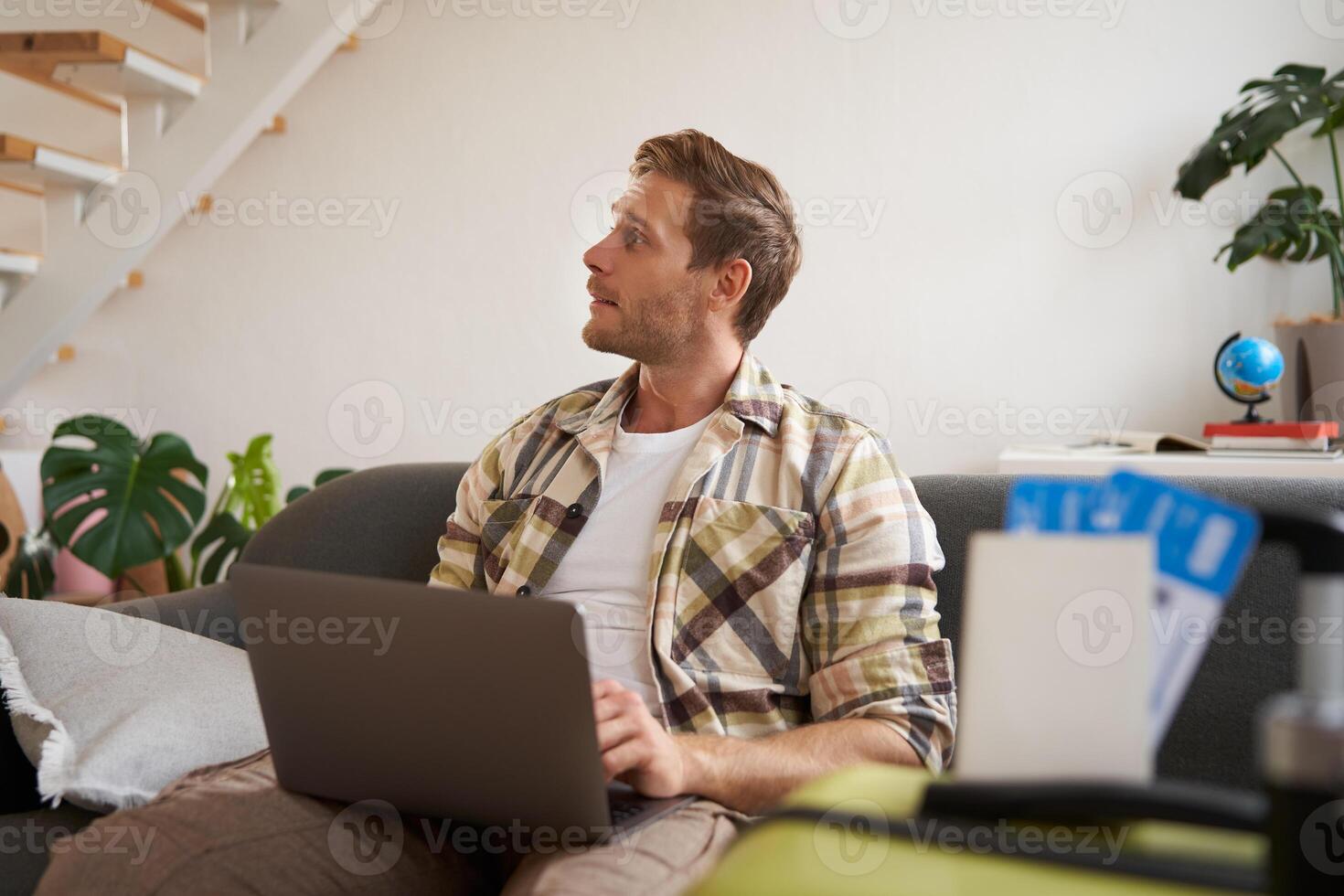 Portrait of young handsome man sitting with laptop, has passport and two plane tickets prepared, planning his journey on computer, booking hotels and restaurants online for the trip photo