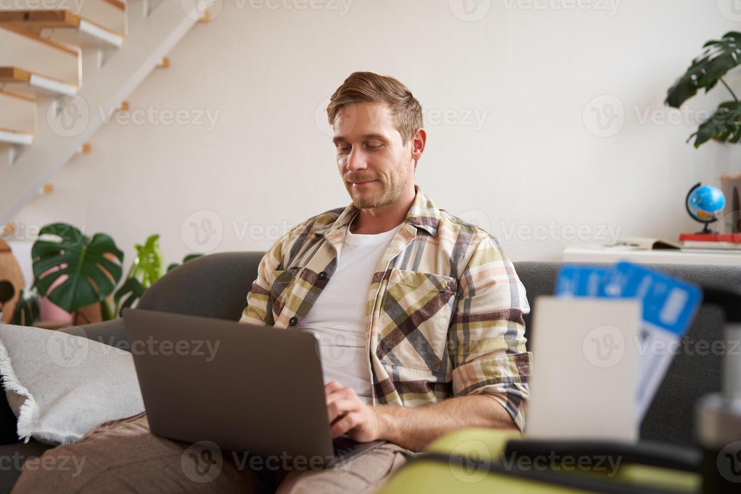 Portrait of handsome young man, has two tickets and passport, goes online on laptop, booking hotel and planning his holiday, sitting in living room photo