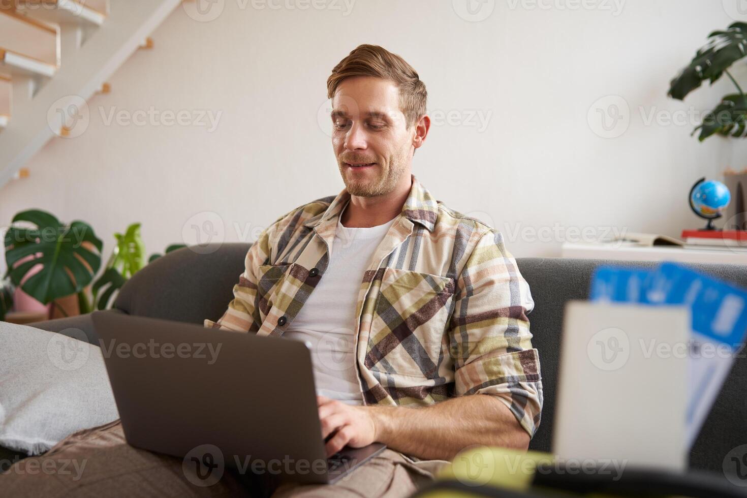 Portrait of handsome young man, has two tickets and passport, goes online on laptop, booking hotel and planning his holiday, sitting in living room photo