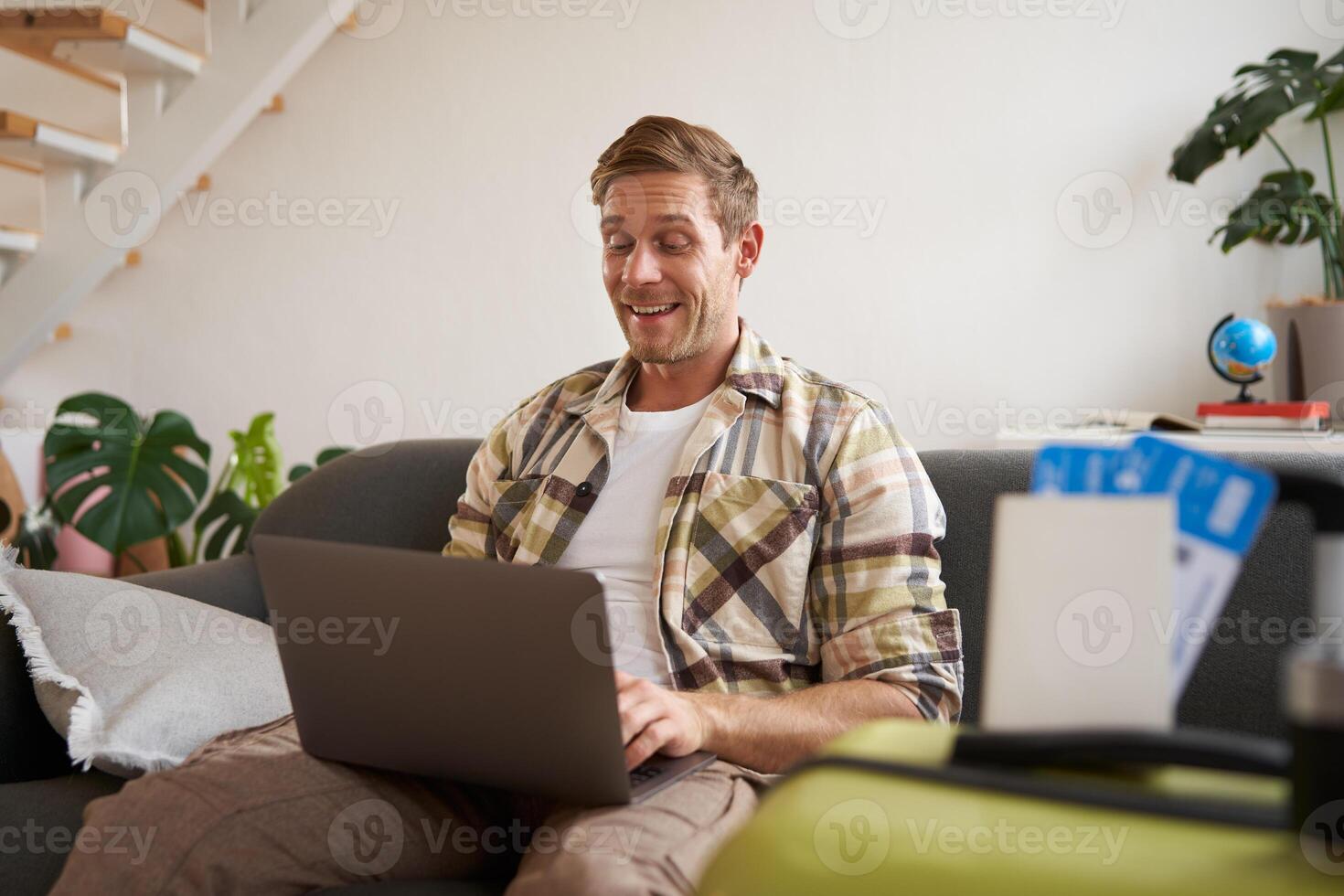 Portrait of happy young man booking holiday for two, has plane tickets, looks at laptop, sits in living room with computer photo