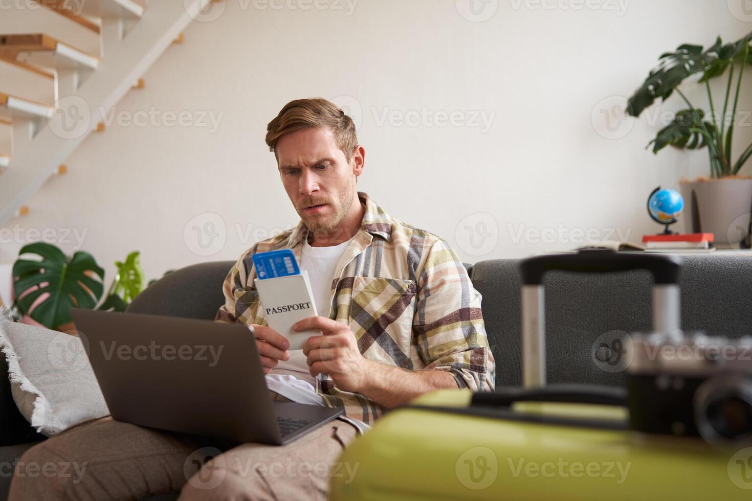 Portrait of grumpy man, holding tickets and passport, has laptop opened, looking concerned and angry, sitting with suitcase on sofa photo