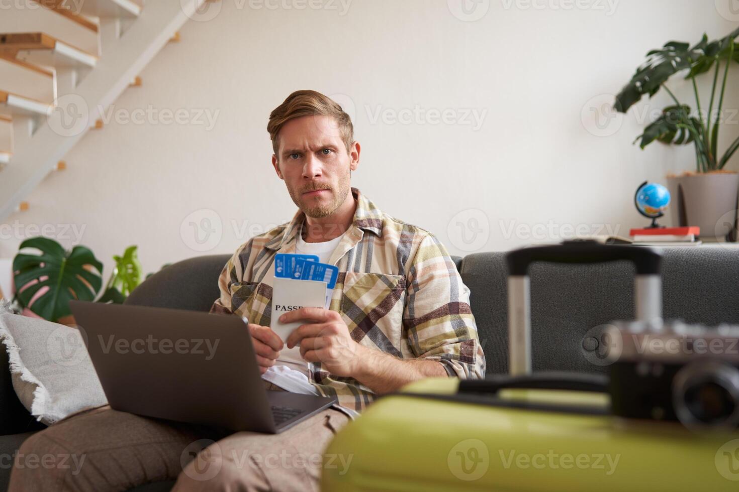 Portrait of serious man with laptop, holds two flight tickets, frowns and stares at camera with concerned, angry face expression photo