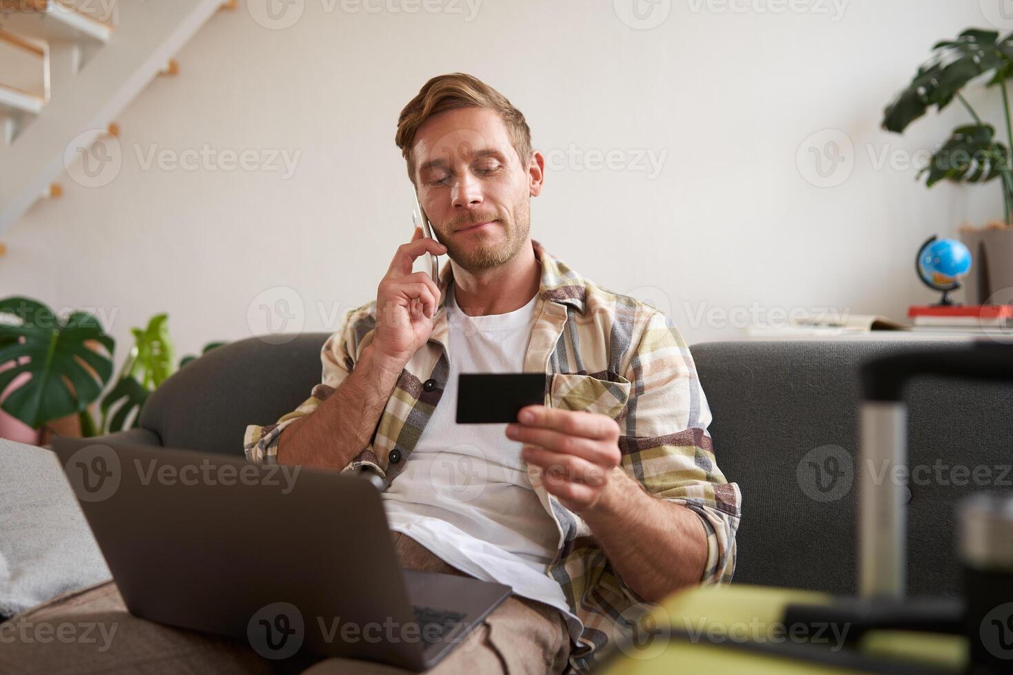 Portrait of young man with credit card, sitting and using laptop, making a phone call, booking hotel or tickets, confirming his purchase over telephone, has suitcase near him photo
